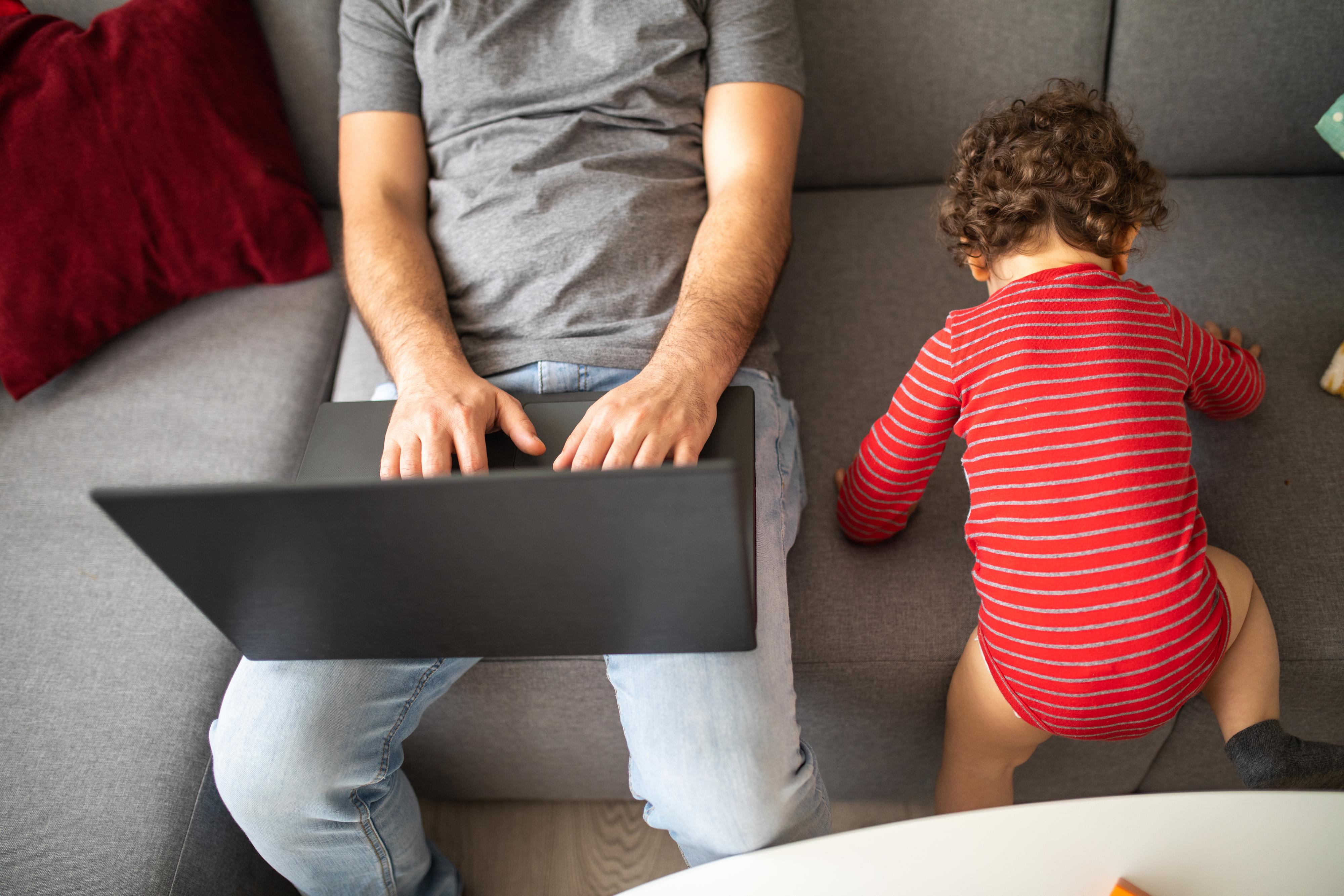 Mies työskentelee kotona kannettavalla tietokoneella, taaperoikäinen lapsi leikkii vieressä sohvalla.